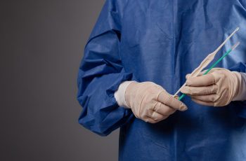 A gynecologist with a venus mirror in his hands. Gynecological accessories, brush, stick. A doctor in a medical mask, a robe, gloves, a hat with a gynecological mirror on a gray background.